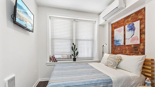 A bedroom with a bed, a window with blinds, a potted plant, and artwork on the wall. - Ututu Stay West (Philadelphia)