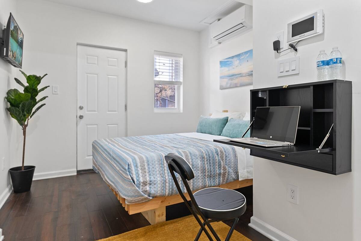 A bedroom with a bed, a desk with a laptop, a chair, a potted plant, a door, and a window.