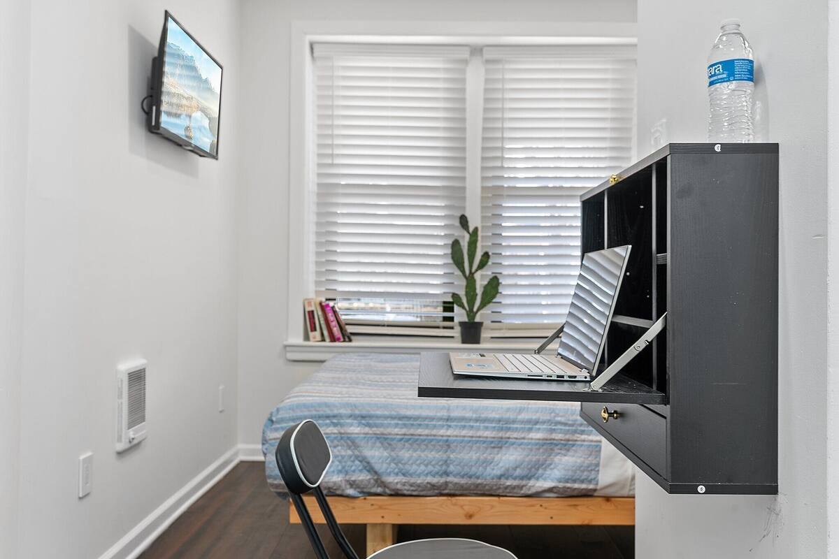A bedroom with a bed, a desk with a laptop, a chair, a window with blinds, a potted plant, and a water bottle on the wall.