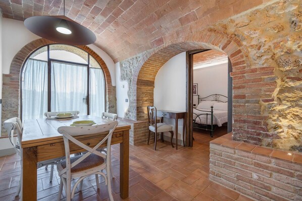 A room with a rustic brick wall, a wooden dining table with chairs, a bed with a metal frame, and a large window with curtains.
