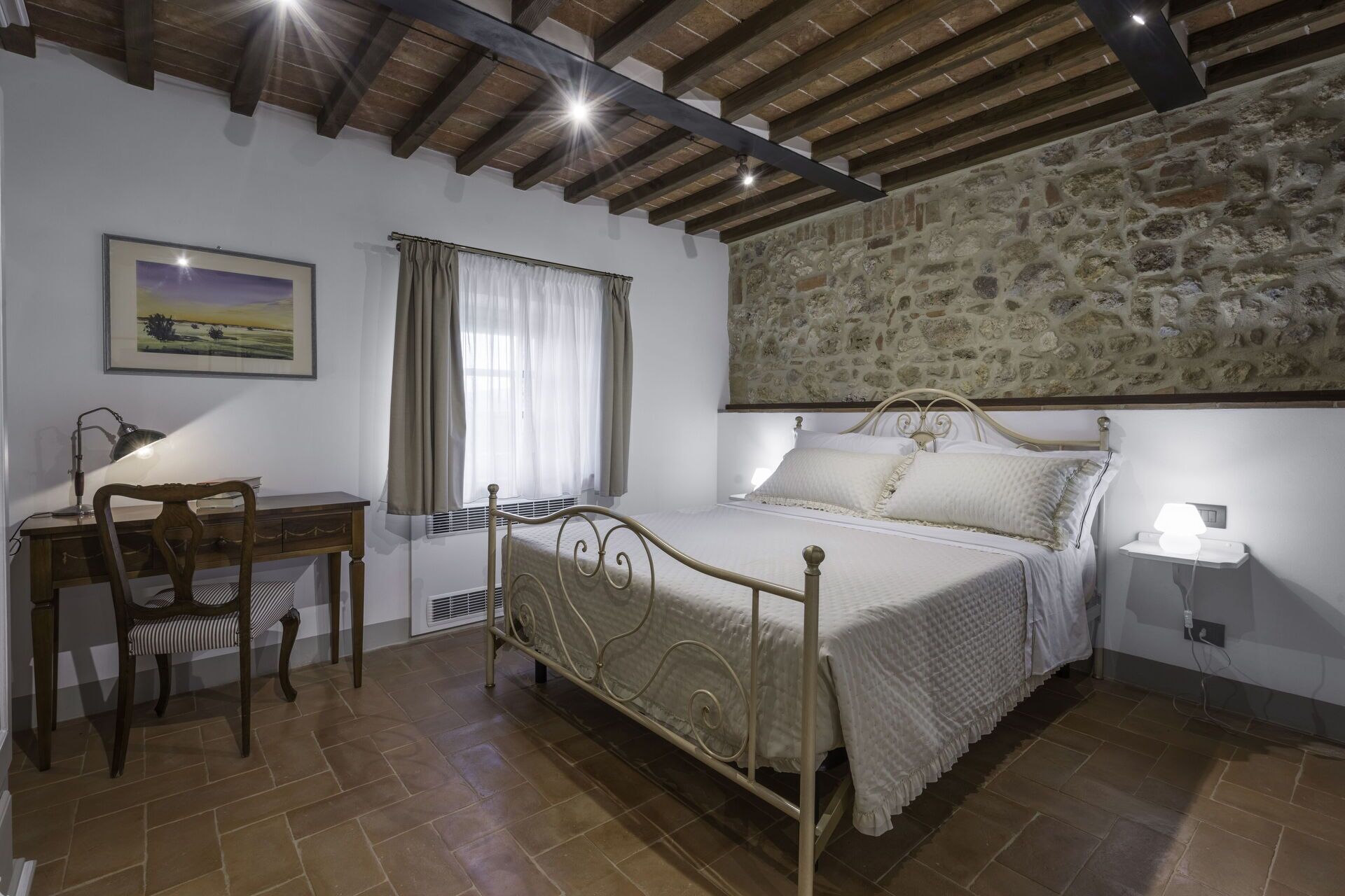 A bedroom with a stone wall, a bed, a desk with a chair, and a window with curtains.
