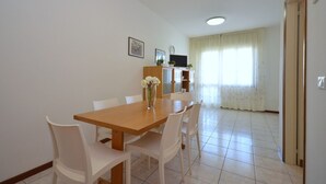 A dining area with a wooden table and white chairs, a vase with flowers, and a kitchen in the background.