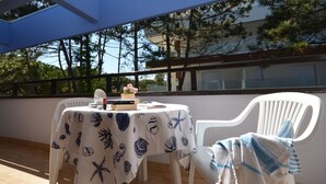 An outdoor patio with a table covered by a tablecloth with a shell pattern, a white plastic chair, and a blue and white striped towel draped over the chair.