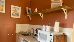 A compact kitchen with a white refrigerator, a microwave, a sink, and a wooden shelf above. - Bedroom 1 - La Rêveuse (PEUPLINGUES)