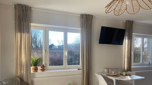 A modern living room with a round wooden table, a white desk, a comfortable armchair, and a TV mounted on the wall. - Bedroom 1 - La Rêveuse (PEUPLINGUES)