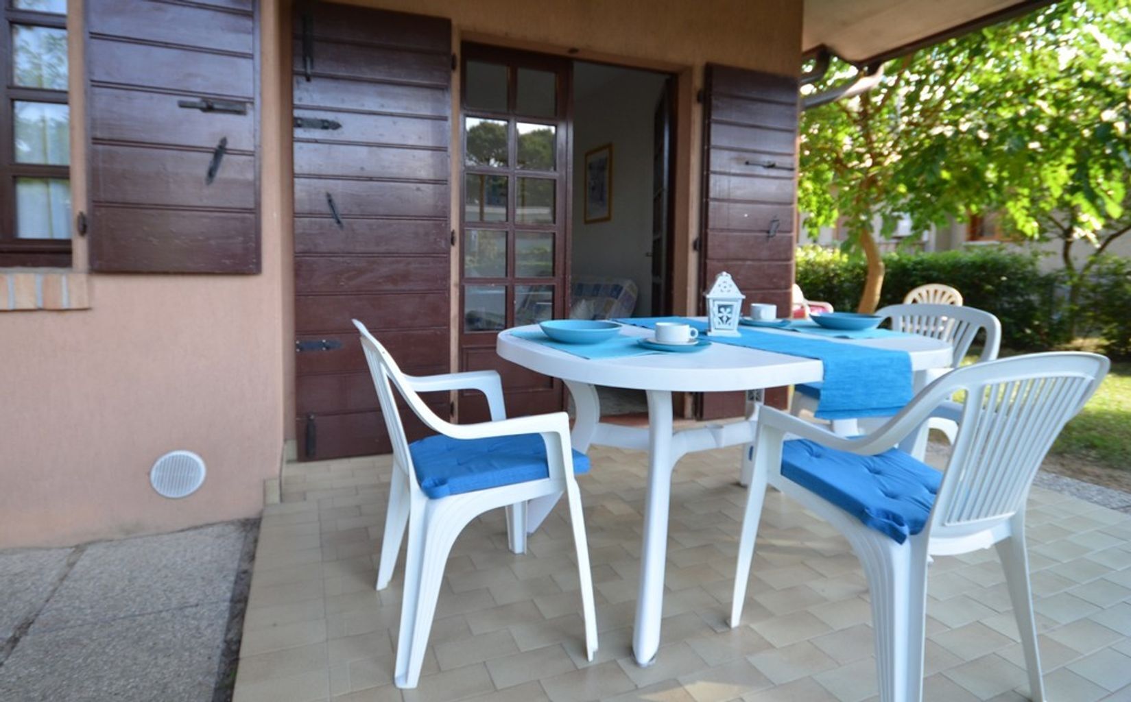 Een terras met een ronde tafel en vier witte stoelen met blauwe kussens, een bruine deur en een raam met zonwering.