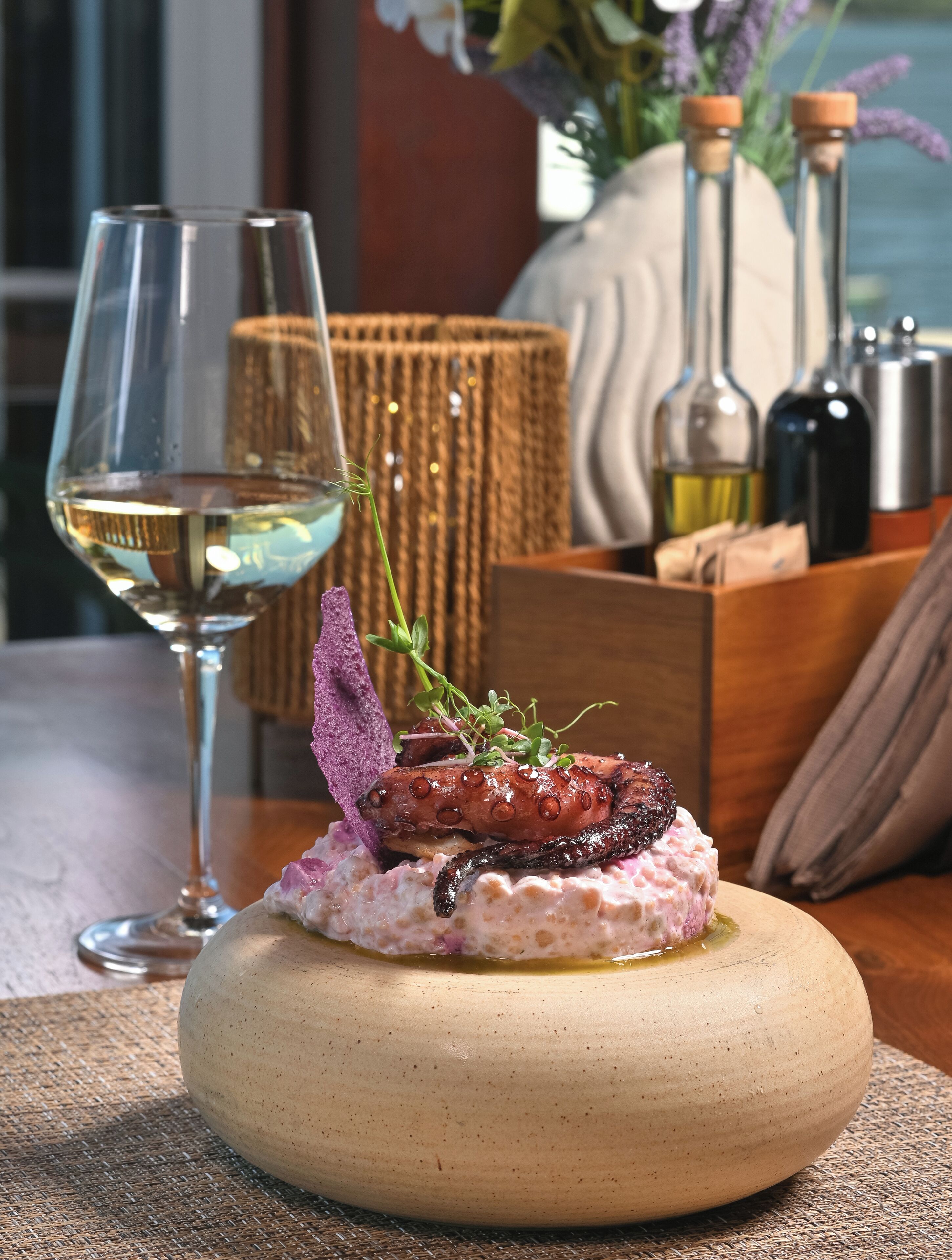 Un plat de poulpe servi dans une assiette ronde en céramique, accompagné d’un verre de vin blanc, sur une table où l’on aperçoit un panier tressé et des bouteilles en arrière-plan.