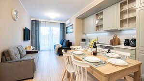 Cozinha moderna com armários brancos, uma mesa de jantar posta para quatro pessoas e uma sala de estar com sofá e televisão.