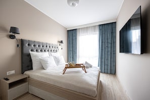 A modern bedroom with a tufted headboard, a bedside table with a remote control, a tray with breakfast items, and a large window with curtains.