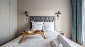 Quarto de hotel moderno com cabeceira estofada, duas luminárias de cabeceira, bandeja de madeira com itens de café da manhã e uma toalha enrolada na cama.
