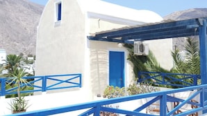 A white building with a blue door and window, surrounded by blue railings and palm trees. - Black Beach Village (Santorini)