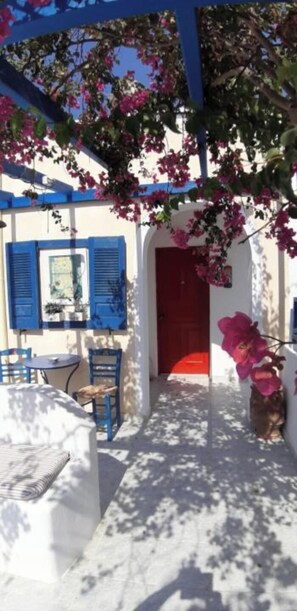 A covered outdoor seating area with a white table and blue chairs, a red door, and pink flowers. - Black Beach Village (Santorini)