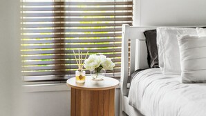 A bedroom with a white bed, a round wooden nightstand, and a window with horizontal blinds.