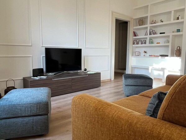A modern living room with a flat-screen TV on a wooden console, two sofas, and a white shelving unit with decorative items.