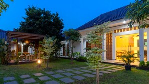 A residential building with a tiled roof, a covered patio with seating, and a well-maintained garden with stepping stones. - WAKAYAPA Syariah Residence (Kelapa Dua)