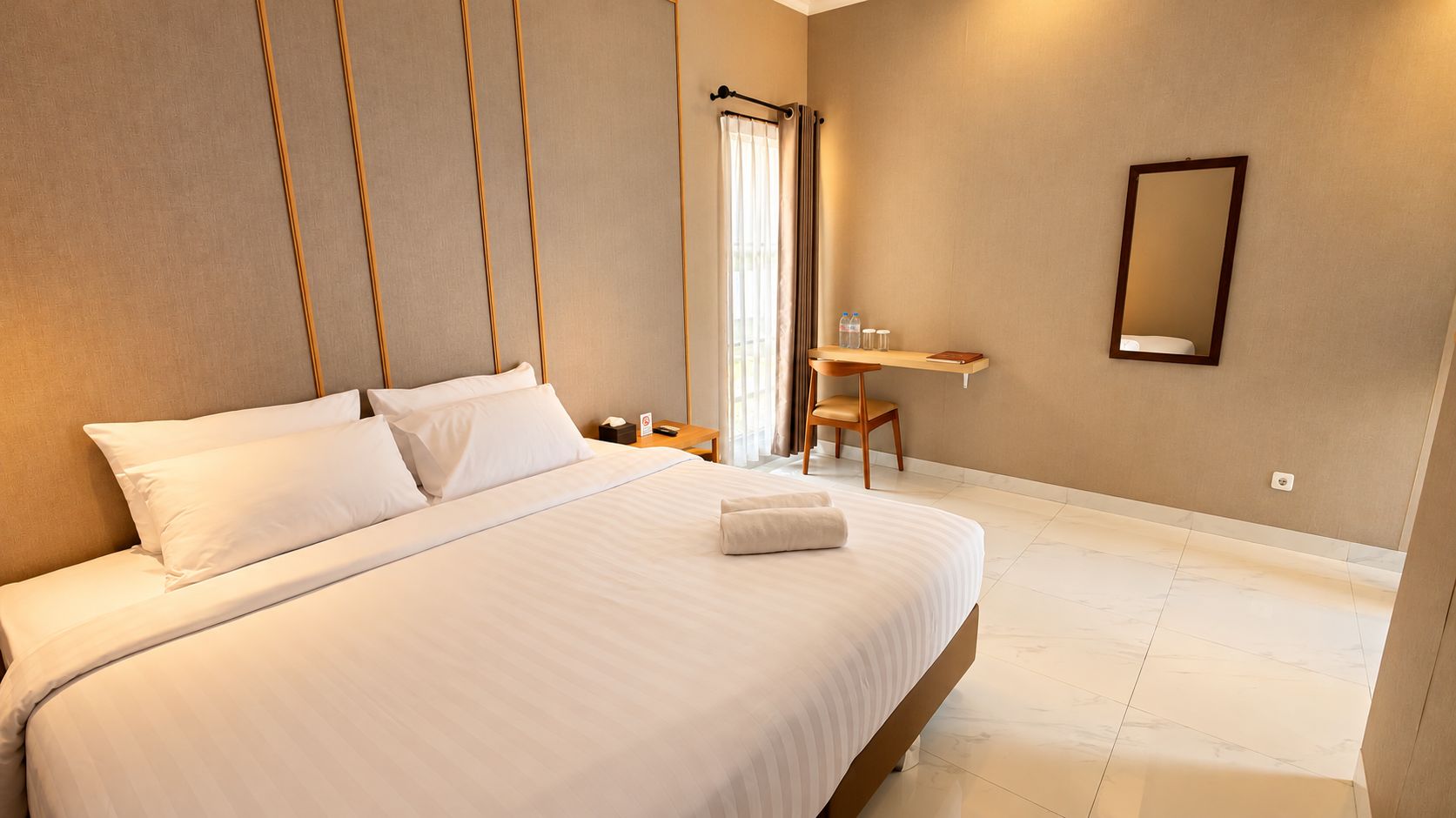 A hotel room with a large bed, a bedside table, a chair, a desk, a mirror, and a window with curtains.