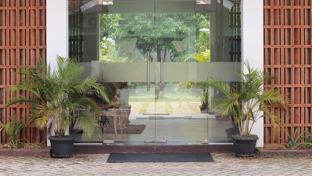 A modern entrance with glass doors, potted plants, and a brick pathway. - WAKAYAPA Syariah Residence (Kelapa Dua)