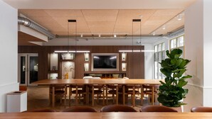 A modern dining area with a long wooden table, multiple chairs, a large flat-screen TV, and a potted plant.
