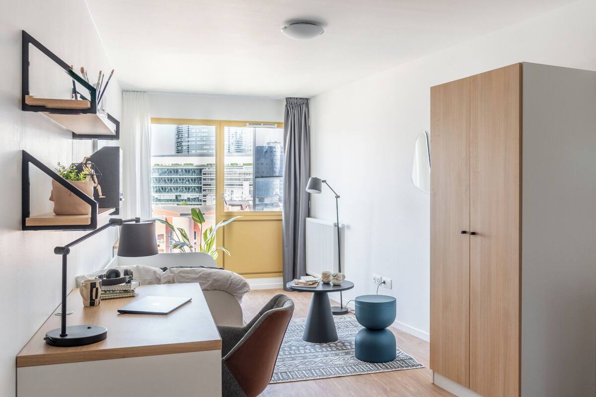 A modern, well-lit bedroom with a large window, a desk with a chair, a wardrobe, and a bedside table with a lamp.