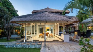A covered outdoor area with a thatched roof, a dining table with chairs, and a stone pathway leading to a house.