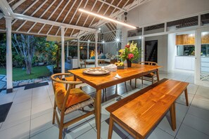 Een ruime eethoek met een houten tafel en banken, omgeven door een glazen constructie met uitzicht op een tuin.