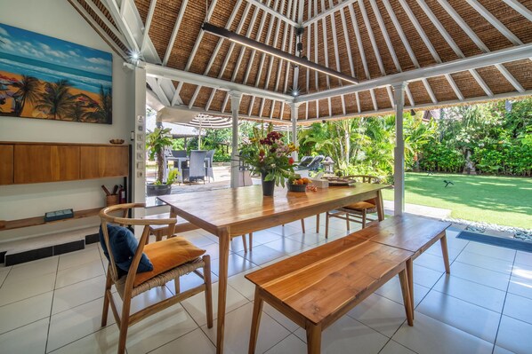 Un patio amplio con una mesa de madera y bancos, rodeado de abundante follaje y vista al océano.