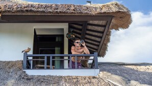 A thatched-roof building with a balcony, a bird perched on the railing, and a person standing on the balcony.