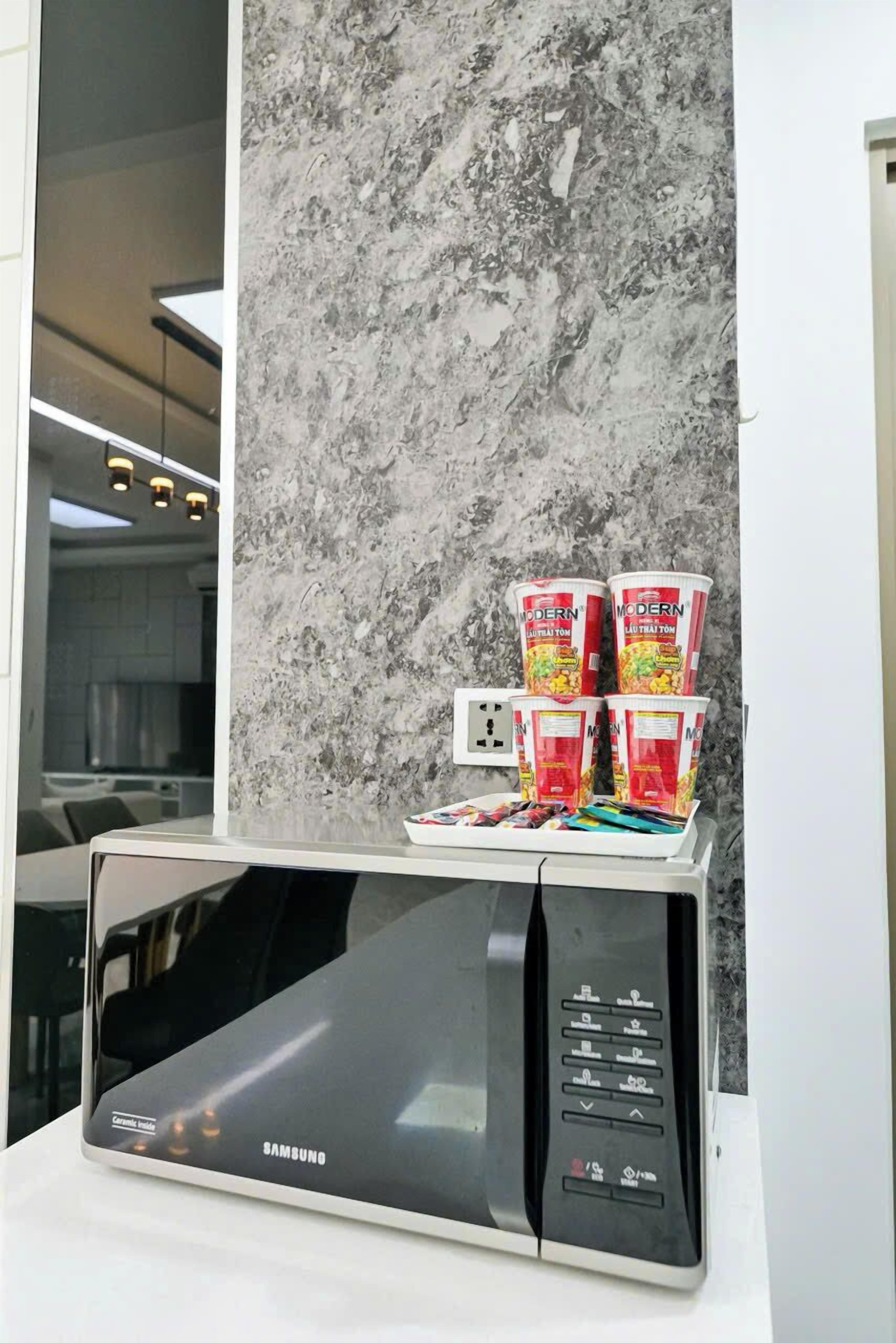 A Samsung microwave on a white countertop with a marble backsplash. There are four Modern brand cans stacked on top of each other.