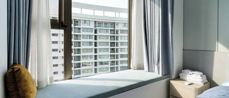 A hotel room with a window seat, a bedside table with towels, and a view of a high-rise building.