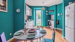 Un cuarto color turquesa con una mesa de comedor lista para cuatro personas, una zona de cocina con gabinetes blancos y una puerta que da a un balcón.