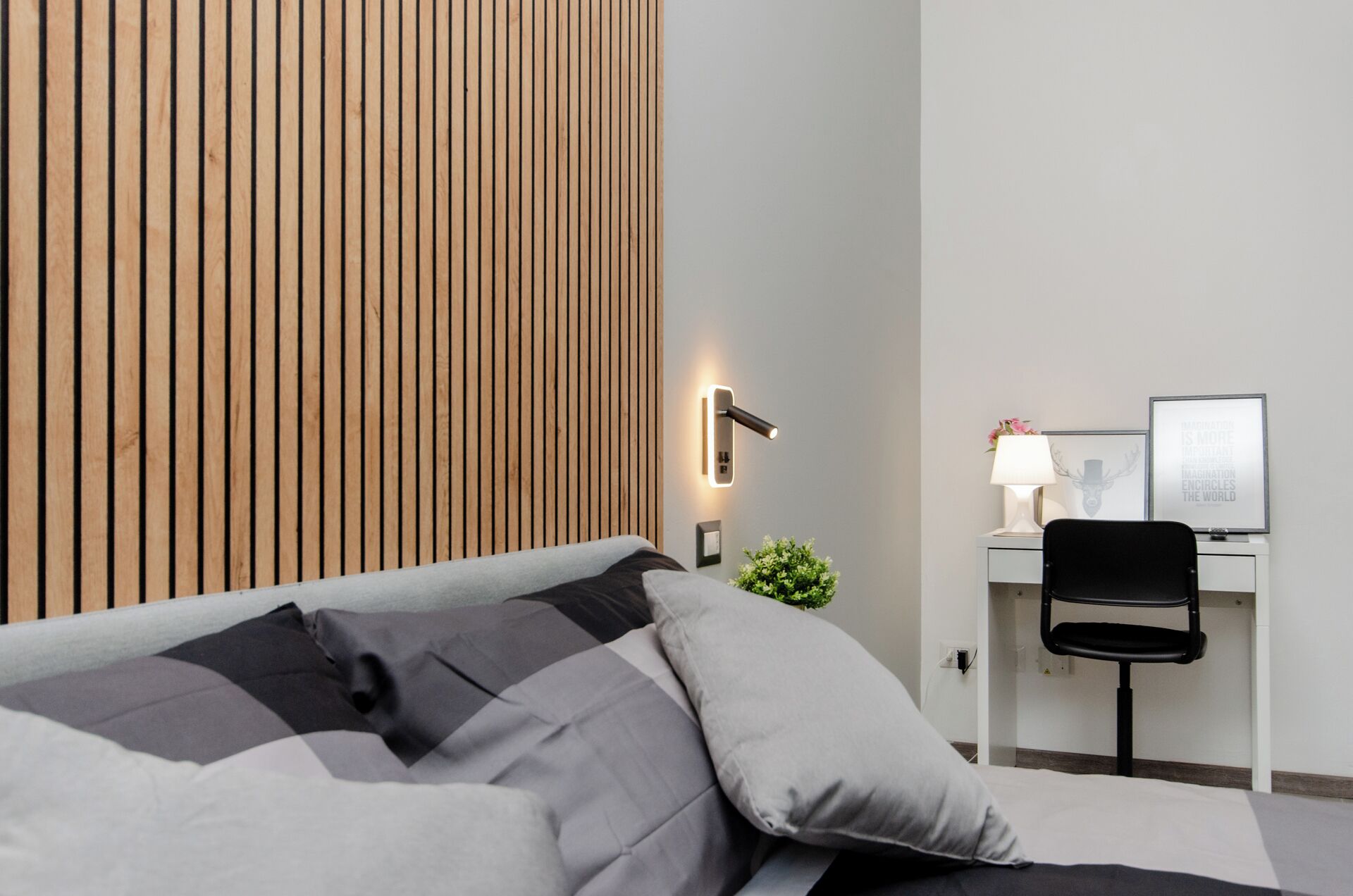 Une chambre d’hôtel moderne avec un mur en lambris de bois, un lit avec une literie grise et noire, un bureau avec une chaise et une petite plante.