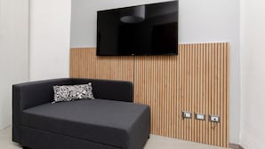 Ein modernes Zimmer mit grauem Sofa, einem schwarz-weißen Kissen, einem Flachbildfernseher, der an einer Wand aus Holzpaneelen befestigt ist, und Steckdosen in der Wand.