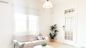 Una habitación luminosa y blanca con un sofá color beige, una mesa redonda de madera y una planta en maceta.