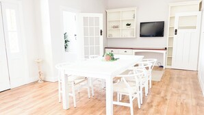 Una habitación luminosa y blanca con suelo de madera, una mesa de comedor blanca con seis sillas, un televisor de pantalla plana montado en la pared y un armario blanco con puertas de cristal.