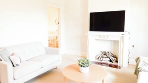 Una sala de estar moderna con un sofá blanco, una mesa de centro redonda de madera con una planta, una chimenea con leña y un televisor de pantalla plana montado encima.