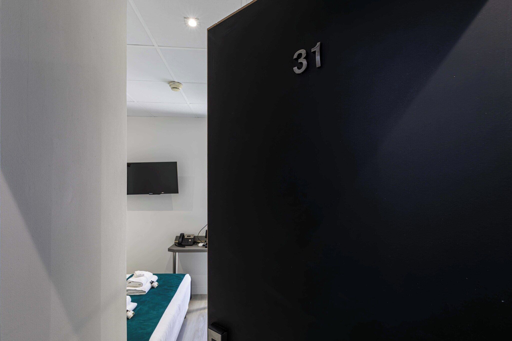 Ein Hotelzimmer mit Bett, Schreibtisch mit Telefon und an der Wand montiertem Fernseher.