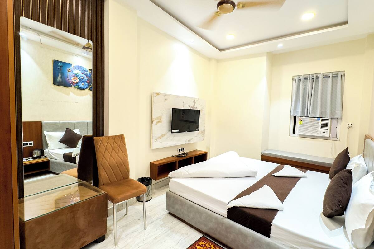 A modern hotel room with a bed, a seating area with a glass table, a television mounted on the wall, and an air conditioning unit.