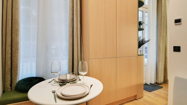 A dining area with a round table set for two, a window with curtains, and a wooden wardrobe.