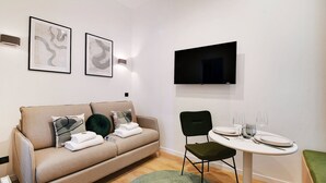 A modern hotel room with a beige sofa, a dining table with chairs, and two framed artworks on the wall.