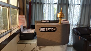 A reception desk with a 'Manager' sign and a 'Reception' sign, a gold lamp, a notice board, and a window with curtains. - Hotel Blue Bird (North Lakhimpur)