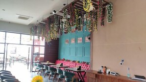 A restaurant with a blue wall, pink cushioned seating, and hanging plants. - Hotel 7b (Chhatarpur)