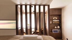 Una habitación de hotel con cama, un separador de madera, un escritorio con silla y una estantería con libros y objetos decorativos.