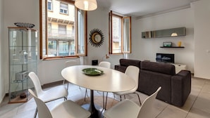 A modern living room with a white dining table, white chairs, a dark brown sofa, and a television on a white cabinet. - Mamo Florence - Santo Stefano Apartment (Florence)