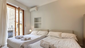 A hotel room with two beds, a window with curtains, an air conditioning unit, and a view of a stone building outside. - Mamo Florence - Santo Stefano Apartment (Florence)