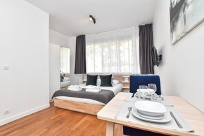 Une chambre d’hôtel moderne avec un plancher en bois, un lit recouvert d’une couette grise et blanche, une table à manger dressée avec des assiettes et des verres, et une grande fenêtre avec des rideaux.