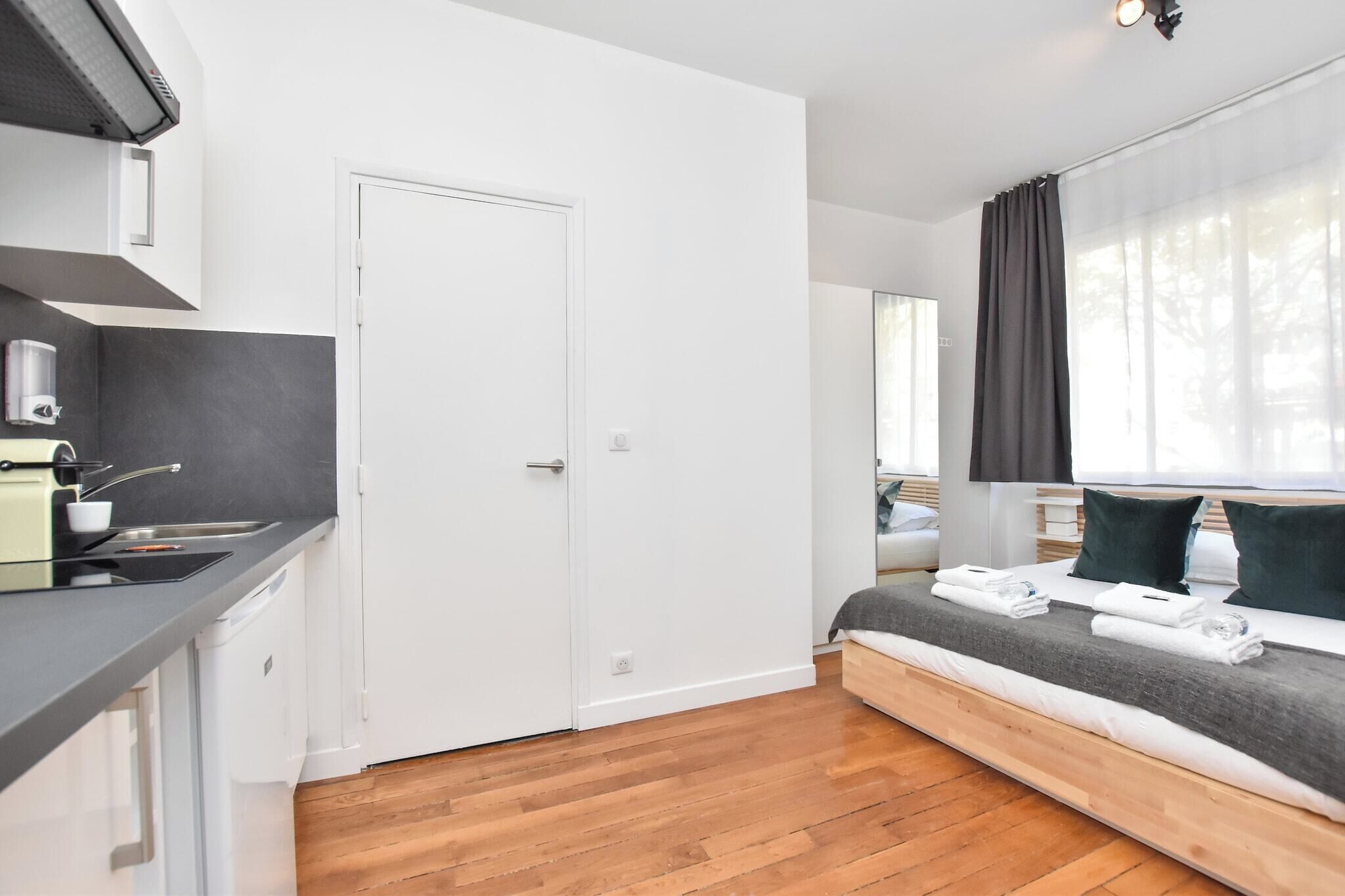 A modern, compact room with a kitchenette, a bed with a grey blanket, and a large window with sheer curtains.