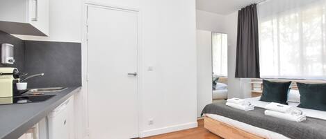A modern, compact room with a kitchenette, a bed with a grey blanket, and a large window with sheer curtains.