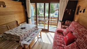A cozy cabin interior with a wooden table and sofa, a sliding glass door to a deck, and a view of a forested area. - Studio 4 persons Châtel / Linga. Chairlift within walking distance. (Châtel)