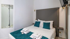A neatly made bed with white linens and teal pillows, a bedside table with a lamp, and a large mirror on the wall.