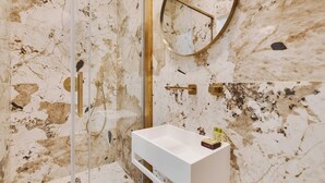 A bathroom with a marble wall, a glass-enclosed shower, a gold showerhead, and a white sink with toiletries.